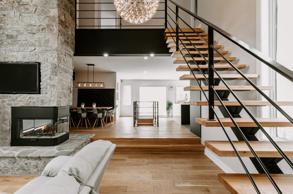 Intérieur de maison moderne avec un escalier en bois et métal noir, un salon avec cheminée en pierre, une salle à manger élégante, et un éclairage design.