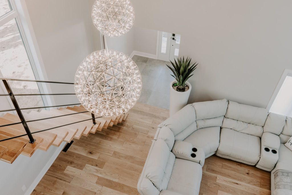 Vue en plongée d'un salon avec un escalier moderne en bois et métal noir, deux suspensions sphériques lumineuses, un canapé en cuir clair, et une plante décorative à côté d'une porte d'entrée.